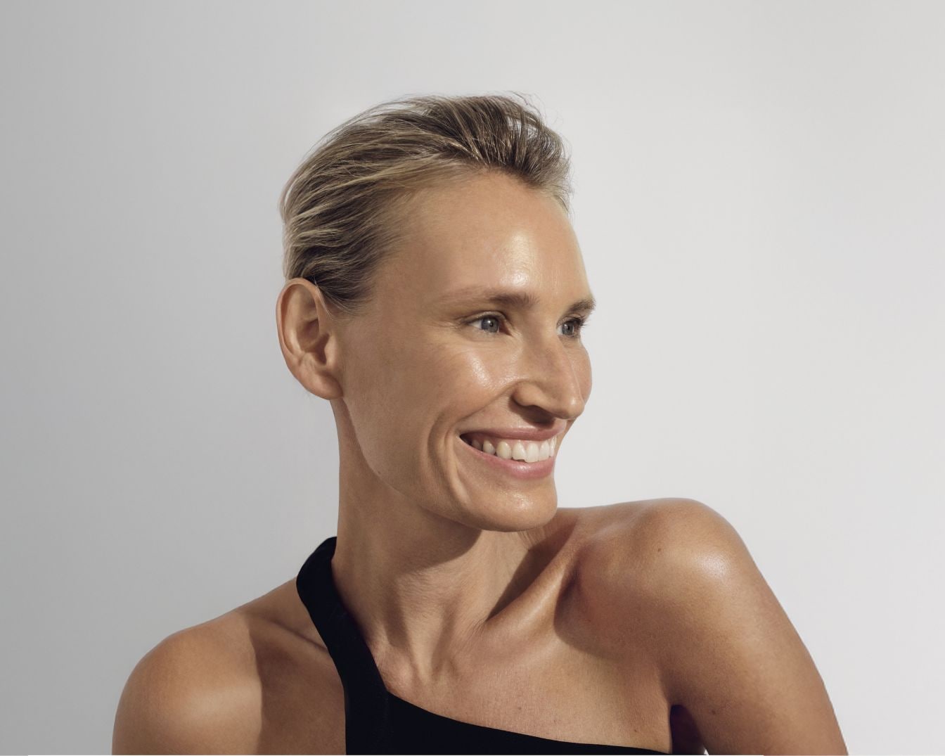 Portret van een lachende vrouw met kort blond haar en een zwarte top, kijkend naar rechts.