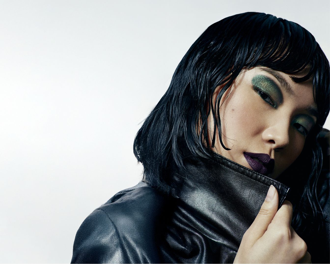 Een vrouw met nat zwart haar, groene oogschaduw en donkerpaarse lippenstift draagt een zwarte leren jas.