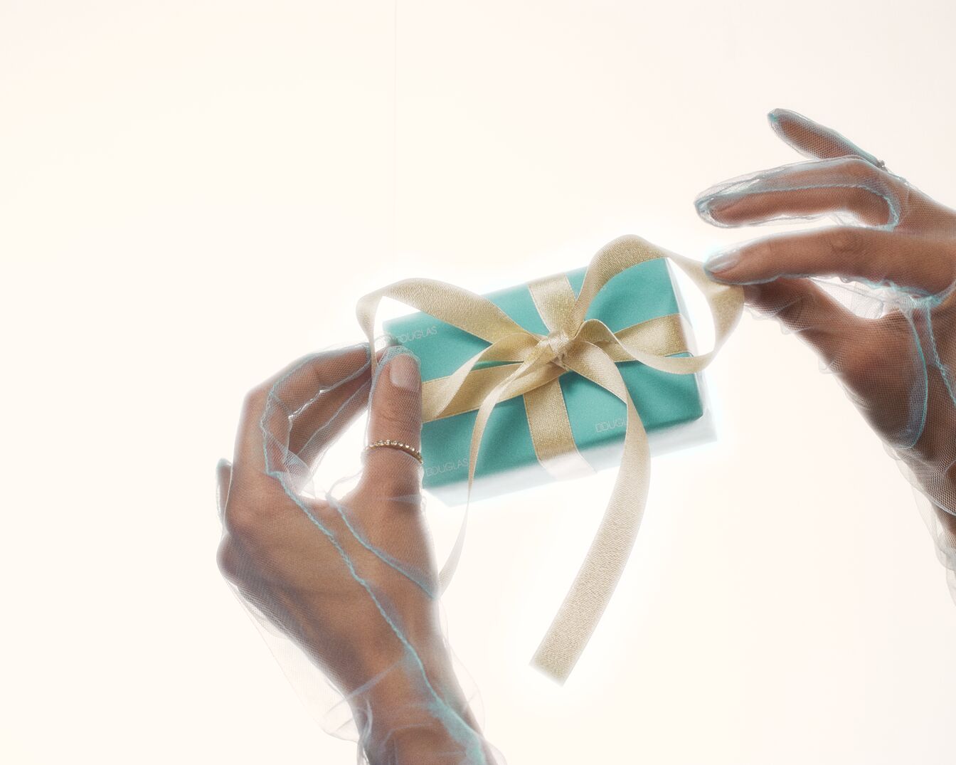 Elegantní tyrkysový dárek od Douglas je převázaný zlatou stuhou a rozvazuje ho ruka v průsvitné rukavici.