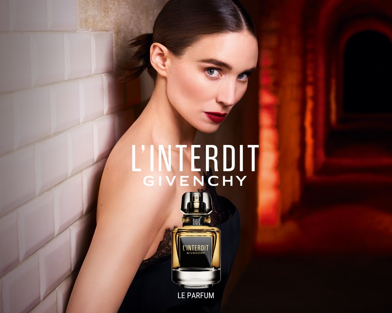 Una mujer apoyada contra una pared de azulejos blancos, con un vestido negro y labios rojos, presenta el perfume L'Interdit de Givenchy en un frasco negro y dorado.
