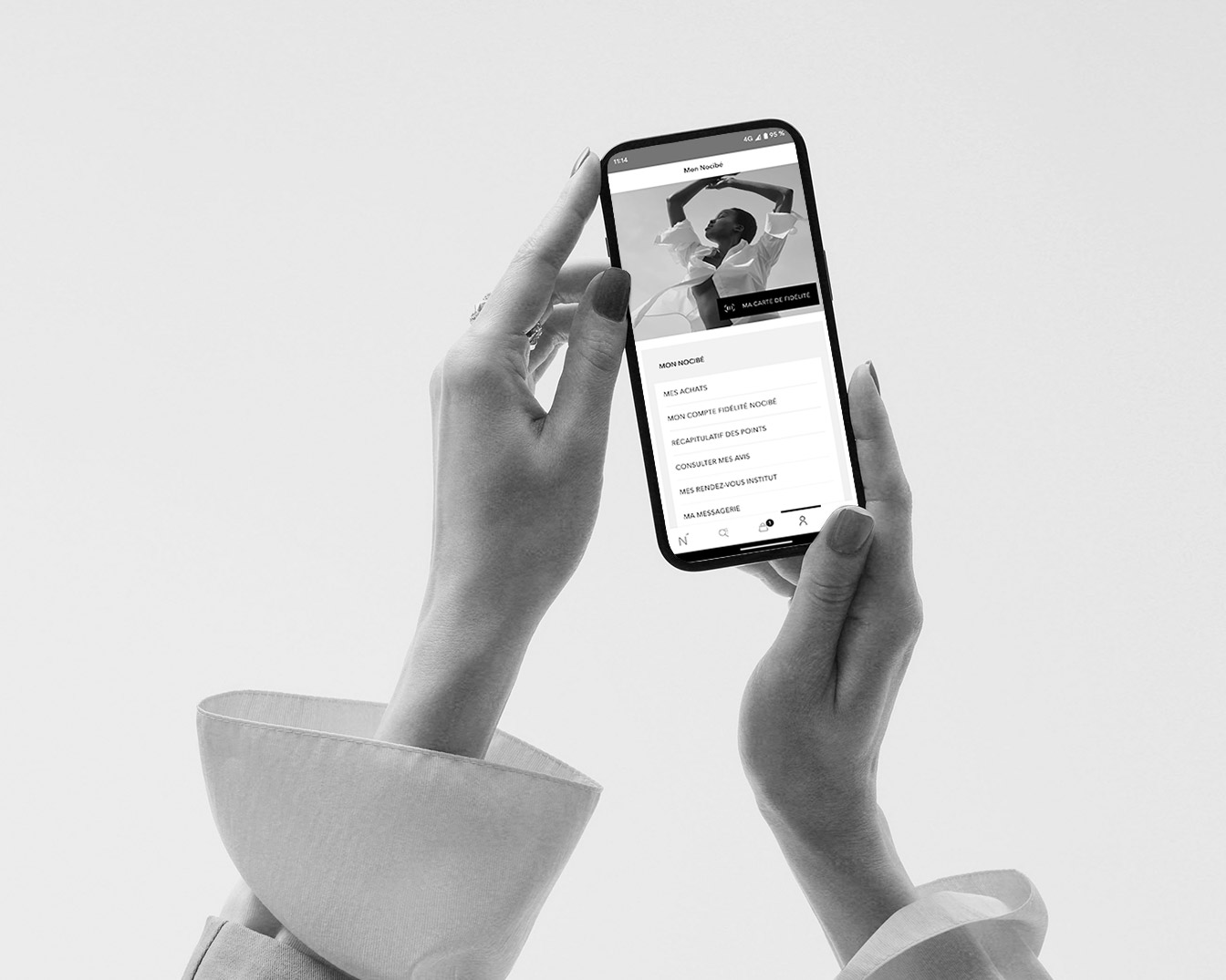 Gros plan sur un smartphone tenu par deux mains manucurées, affichant l'application Nocibé avec les options Mon Nocibé, Mes achats, Mon compte fidélité Nocibé, et plus.