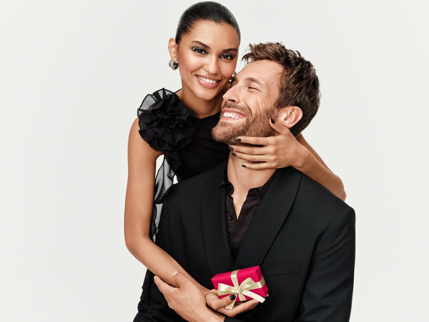 Un couple souriant, l'homme en costume noir tenant un petit cadeau rouge avec un ruban doré, la femme en robe noire avec une fleur sur l'épaule, les mains de la femme autour du cou de l'homme.