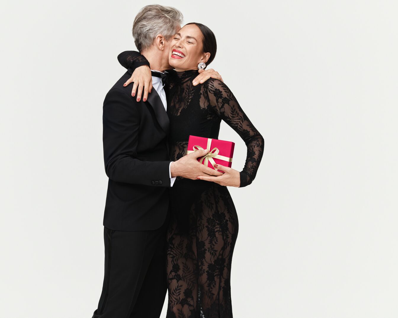 Un homme en smoking noir embrasse une femme en robe de dentelle noire tenant un cadeau rouge avec un ruban doré, rayonnant de joie.