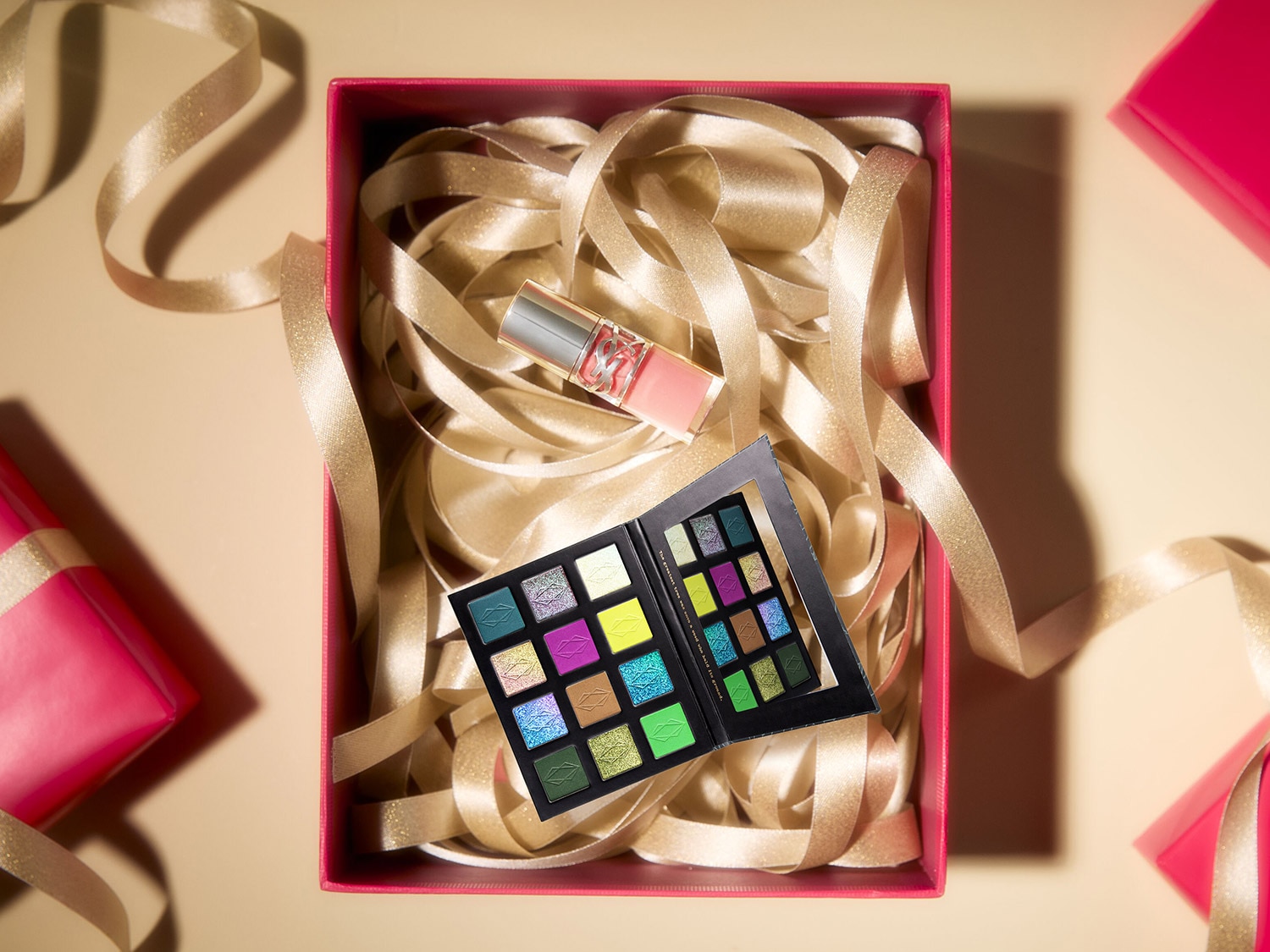 Dans une boîte rose remplie de rubans dorés, on trouve une palette de fards à paupières aux couleurs vives et un brillant à lèvres rose.