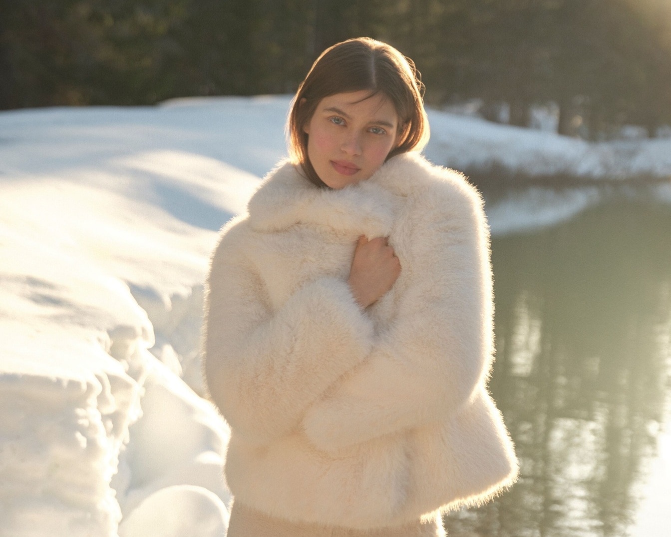 Šviesiame fone jauna moteris, apsivilkusi kreminės spalvos kailinius, švelniai apglėbia save rankomis, spinduliuodama jaukumą ir šilumą.