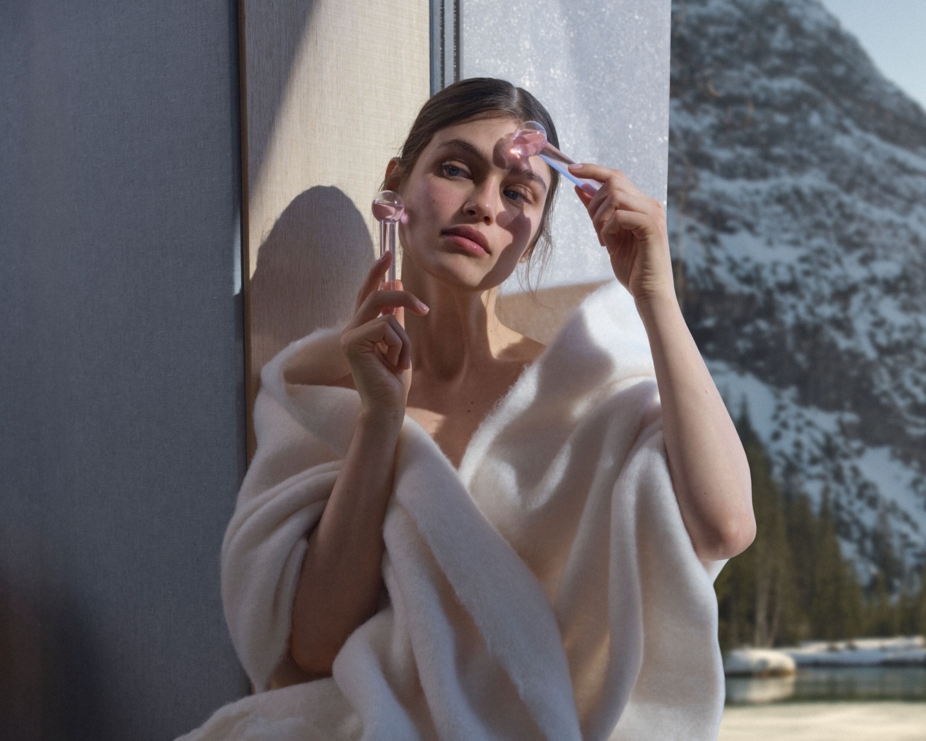 Moteris, apsigobusi baltu chalatu, laiko du rožinius veido masažuoklius, fone matosi snieguoti kalnai, sukuriančius raminančią ir atpalaiduojančią atmosferą.