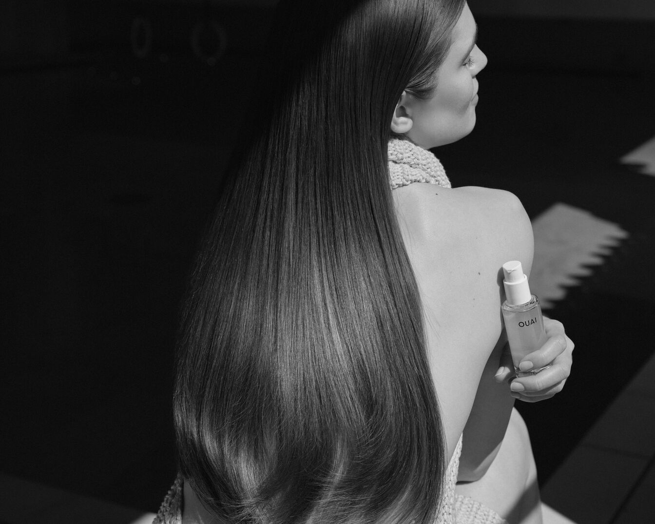 Een zwart-wit foto van een vrouw met lang, steil haar die een flesje QUAI-haarolie vasthoudt, wat de glans en gezondheid van haar haar benadrukt.