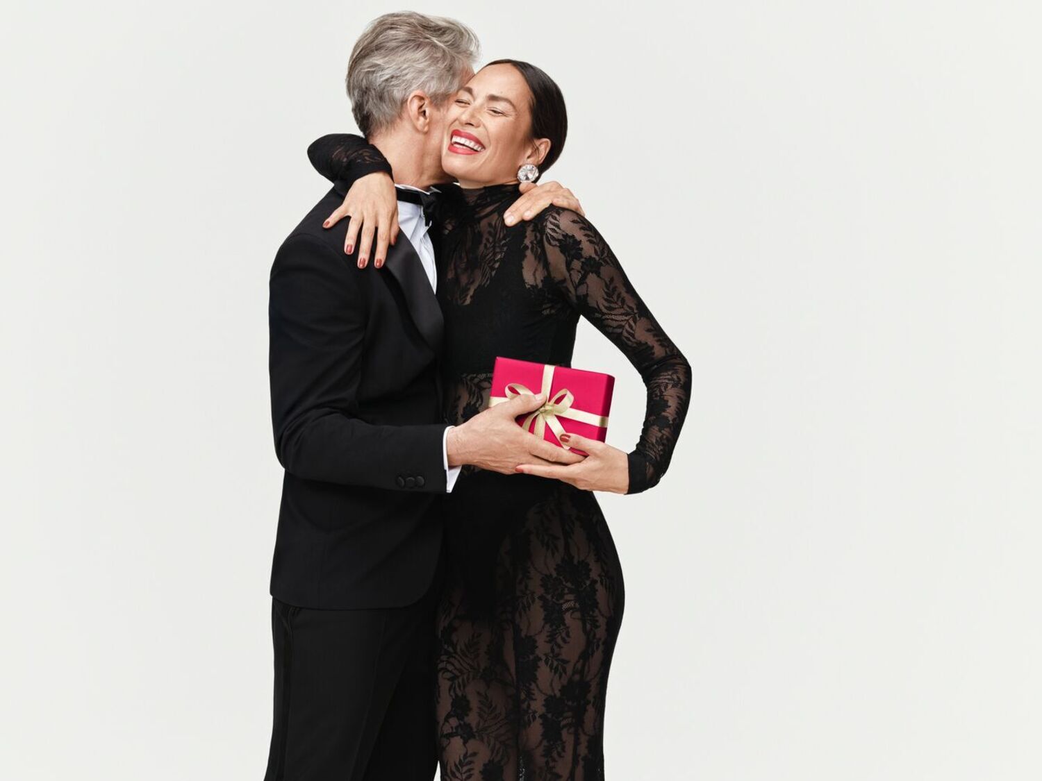 Un homme en smoking embrasse une femme en robe noire en dentelle, tenant un cadeau rouge avec un ruban doré, rayonnant de joie.