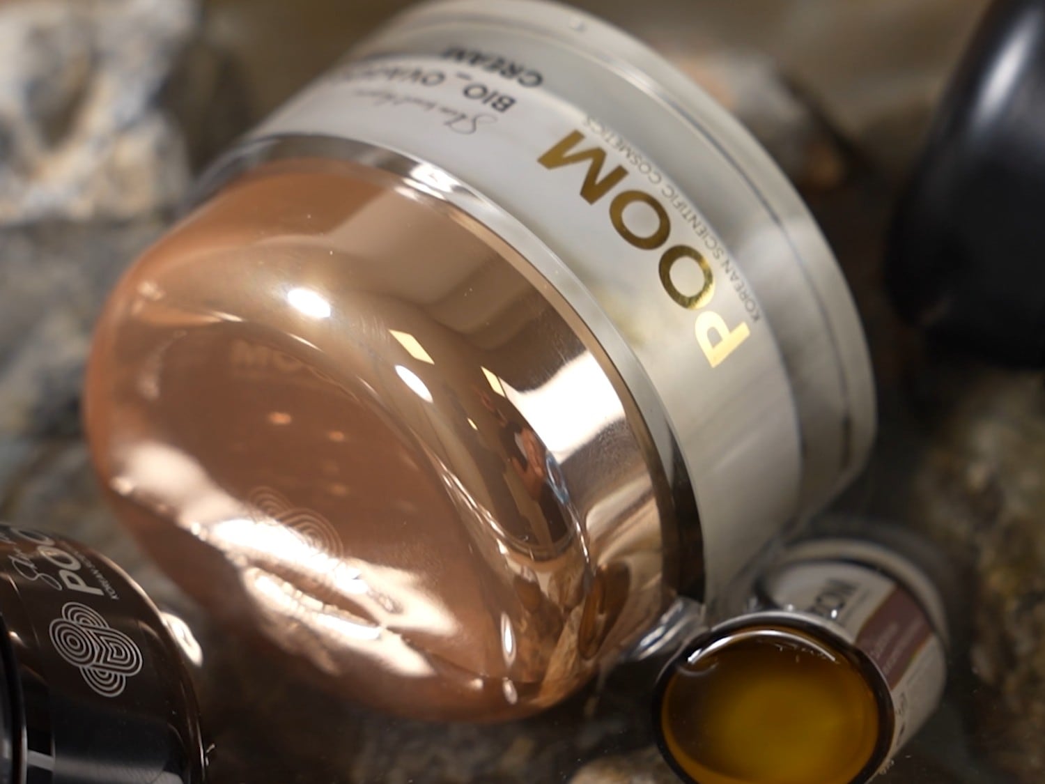 Aquí se muestra un tarro de crema facial POOM Bio Ovine, con un envase dorado brillante y una etiqueta blanca con letras doradas, sobre una superficie rocosa.