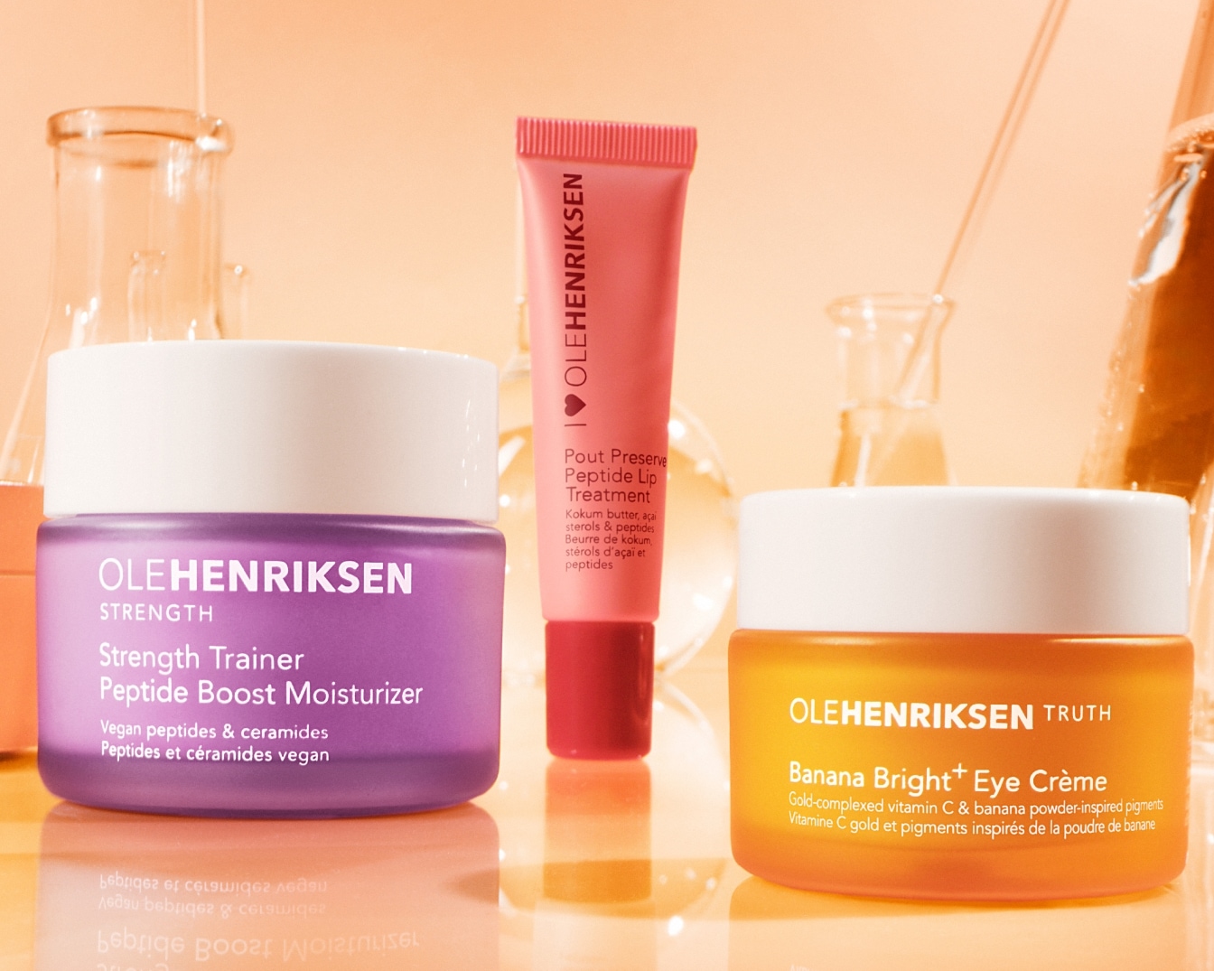 Olehenriksen Strength Trainer Peptide Boost Moisturizer w fioletowym słoiczku, obok Pout Preserve Peptide Lip Treatment w różowej tubce i Banana Bright+ Eye Crème w pomarańczowym słoiczku.