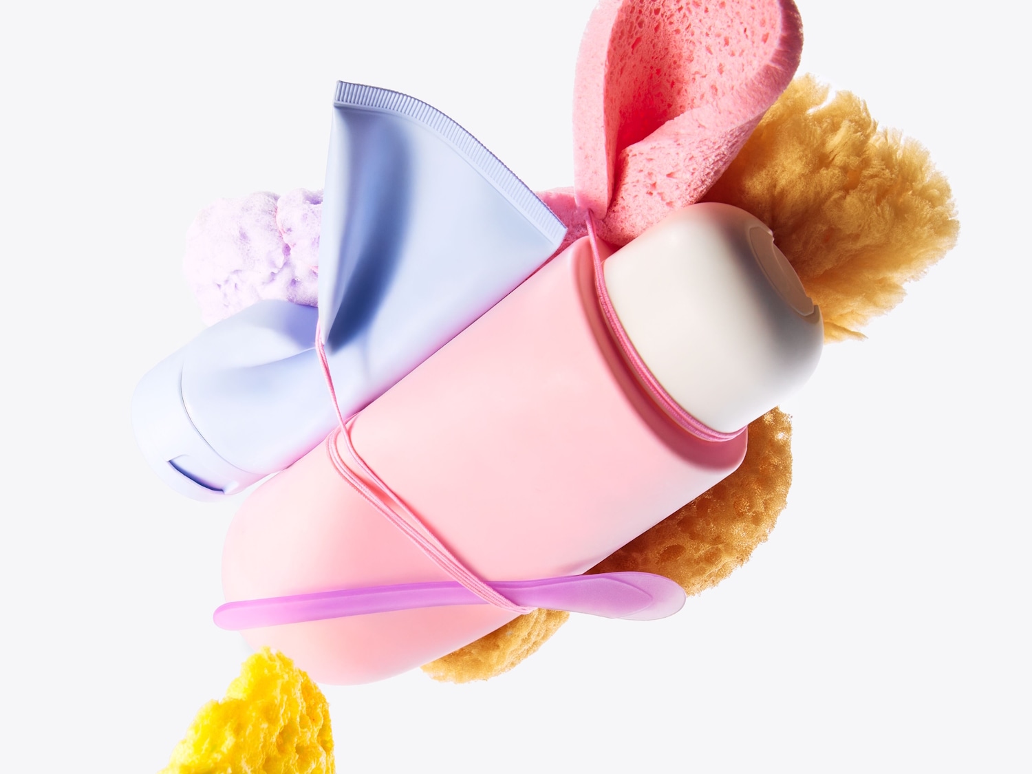 Verschiedene Bad- und Schönheitsprodukte, darunter eine hellblaue Tube, eine rosa Flasche und verschiedene Schwämme in Rosa- und Gelbtönen, die mit einem rosa Band zusammengehalten werden.