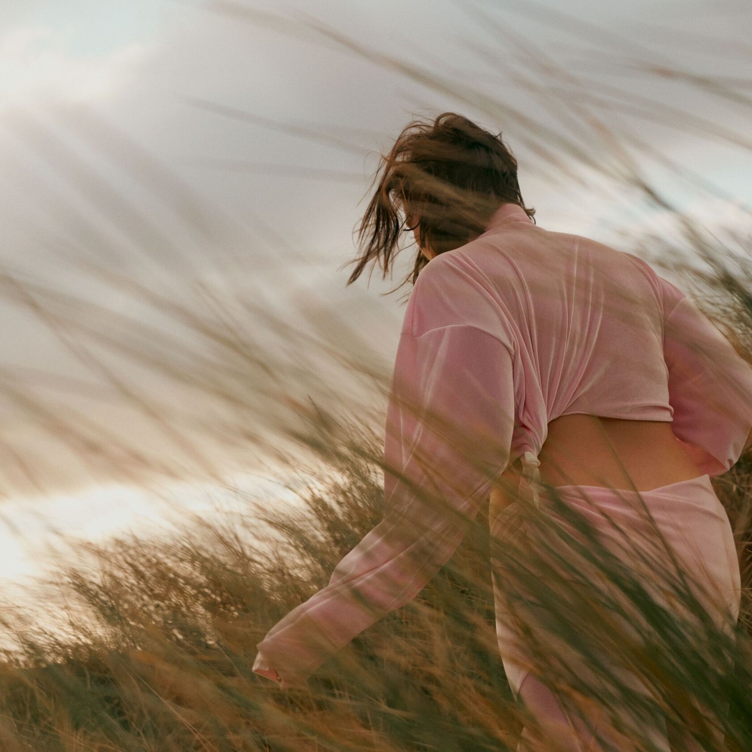 Une femme, vêtue d'un ensemble rose, se promène dans les herbes hautes, dos à la caméra, avec une lumière douce qui illumine la scène.