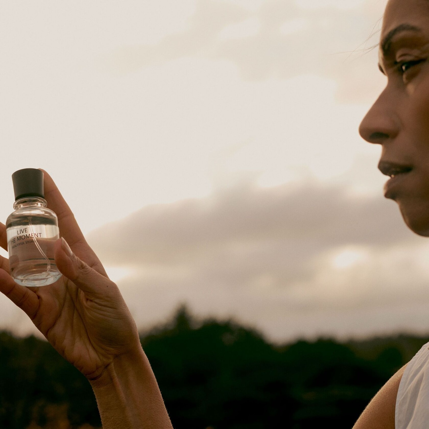 Une femme tient un flacon transparent de parfum "Live the Moment Beautiful Story" avec un bouchon noir, sur un fond de ciel nuageux au coucher du soleil.