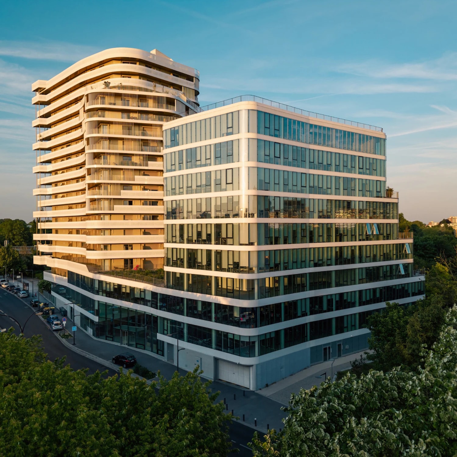 Voici une vue aérienne de deux bâtiments modernes, l'un avec une façade incurvée et l'autre avec une façade en verre rectiligne, sous un ciel bleu clair.