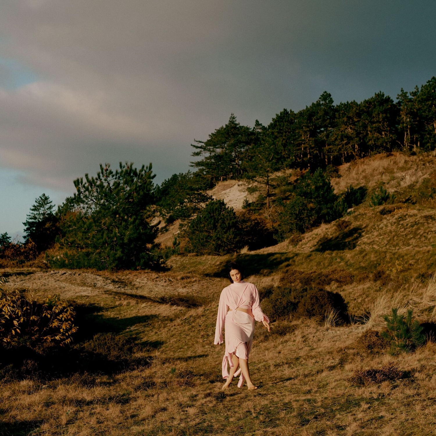 Une femme en robe rose clair se promène pieds nus sur une colline herbeuse, avec des arbres en arrière-plan sous un ciel nuageux.