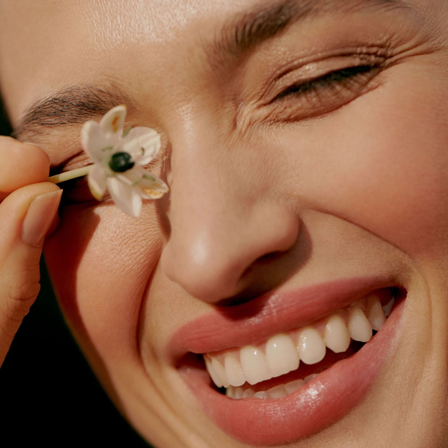 Gros plan d'une femme souriante avec une petite fleur blanche à l'œil, peau éclatante et lèvres roses brillantes.