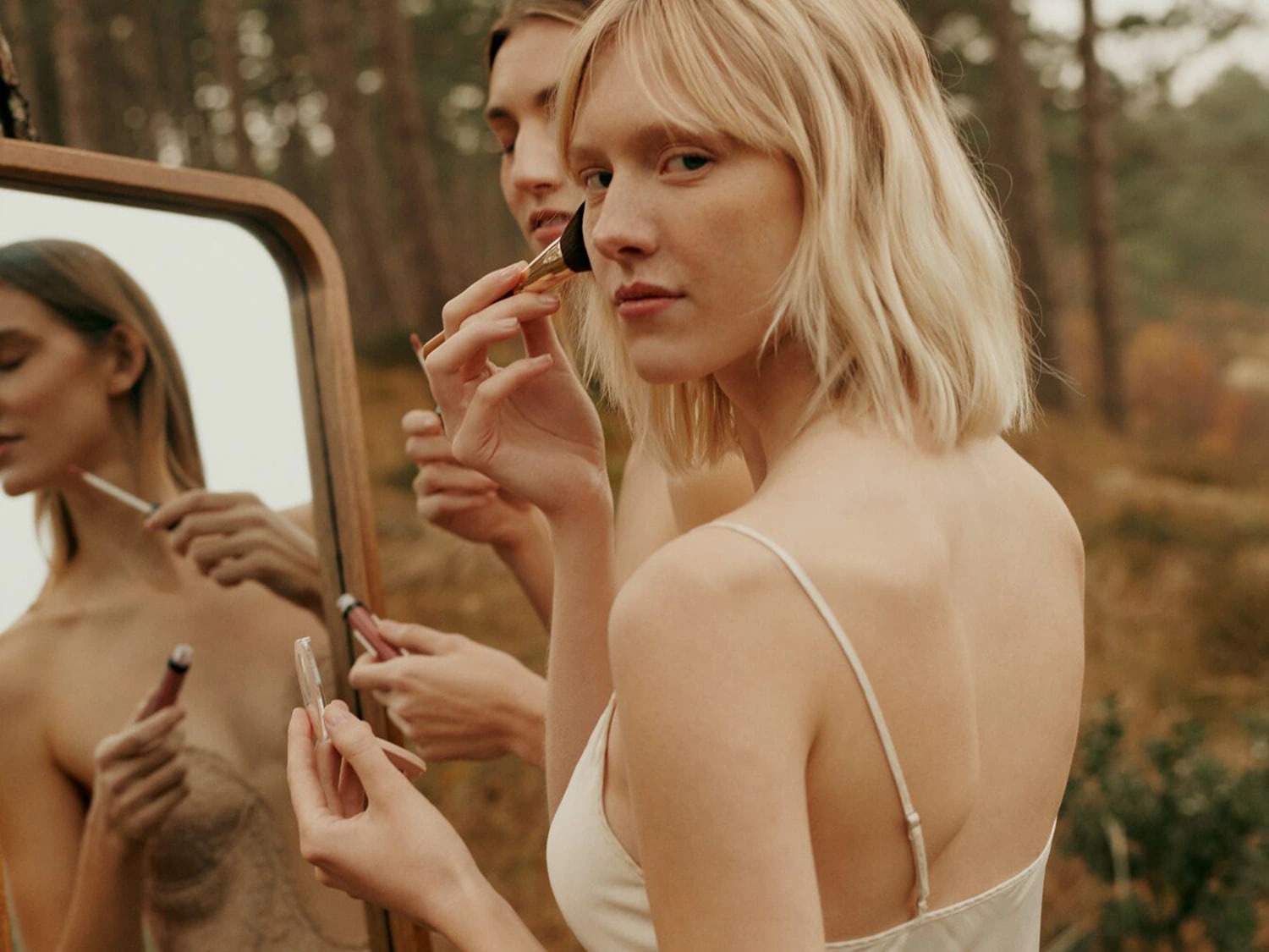 Dans un cadre naturel, une femme blonde applique délicatement du blush avec un pinceau doré, tandis qu'une autre utilise un gloss rosé devant un miroir.