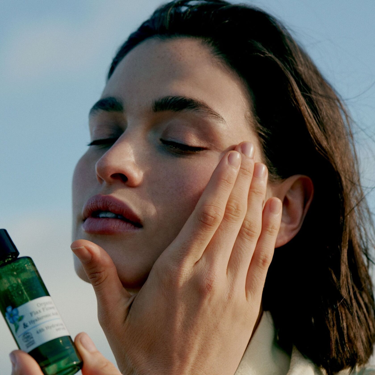 Une femme les yeux fermés applique une lotion hydratante biologique à la fleur de lin dans un flacon vert.