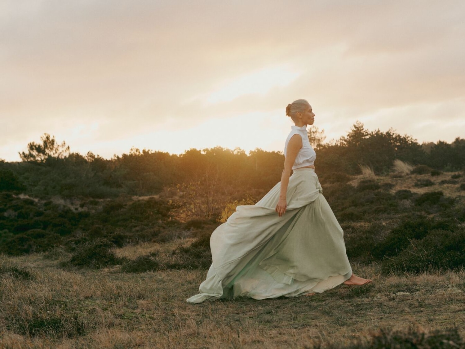 Une femme aux cheveux gris, vêtue d'un haut blanc et d'une longue jupe fluide vert clair, marche pieds nus dans un champ au coucher du soleil.