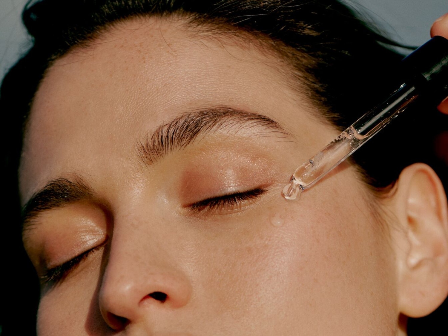 Une femme applique un sérum transparent sur sa peau à l'aide d'une pipette en verre, les yeux fermés, pour un soin ciblé et une peau éclatante.