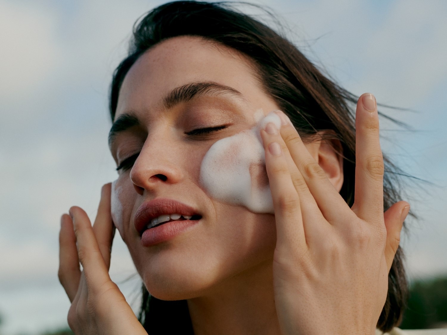 Une femme se nettoie le visage avec une mousse nettoyante blanche, les yeux fermés, pour une peau fraîche et éclatante.