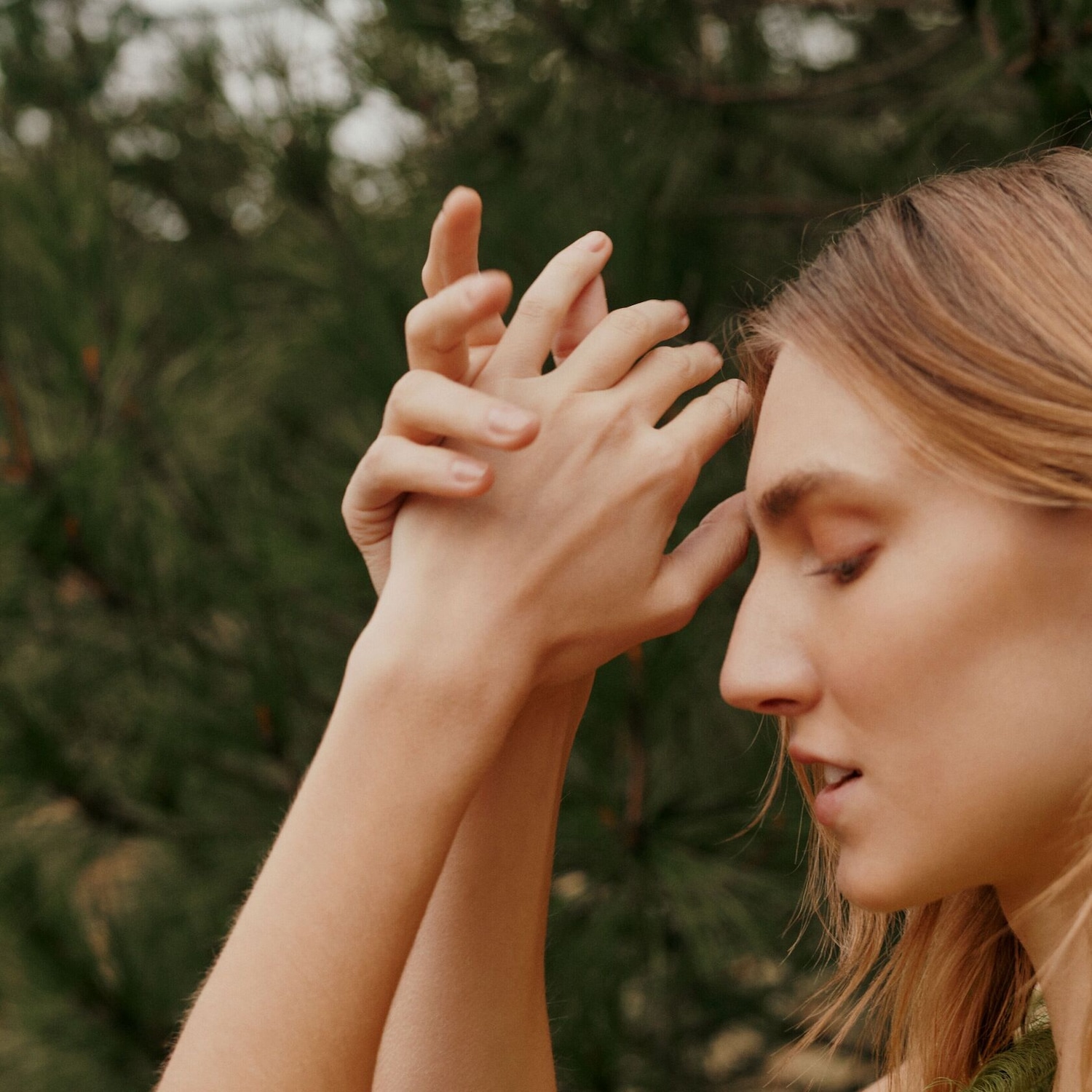 Portrait d'une femme blonde, les yeux fermés, les mains jointes devant son visage, évoquant un moment de calme et de sérénité dans un cadre naturel verdoyant.