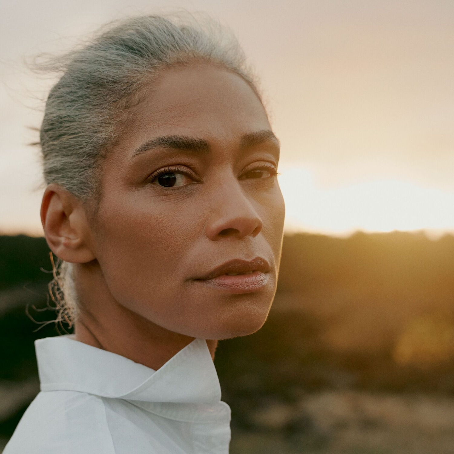Portrait d'une femme aux cheveux gris courts, portant une chemise blanche, regardant intensément vers l'avant sur un fond de paysage ensoleillé.