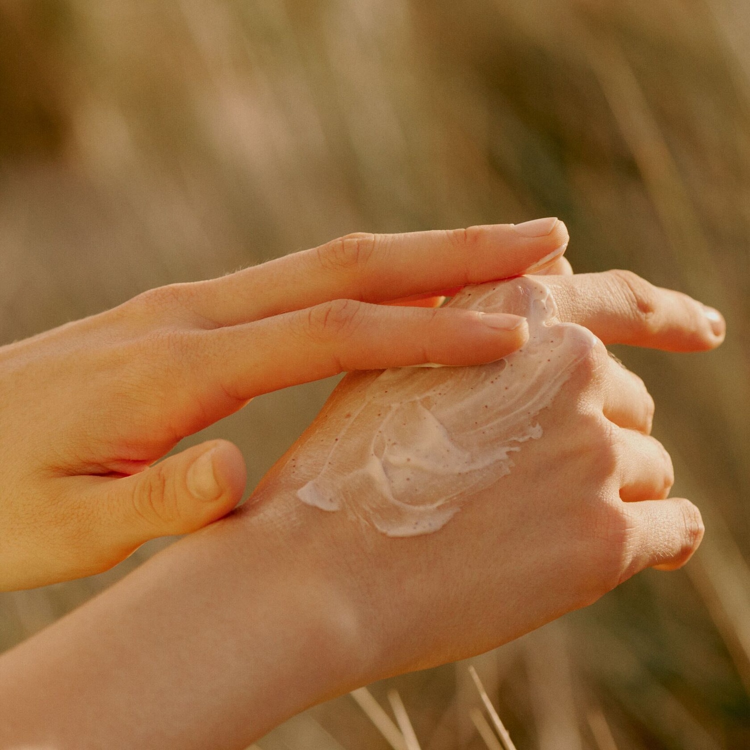 Une main applique une crème hydratante beige sur l'autre main, avec un arrière-plan flou de tons chauds.