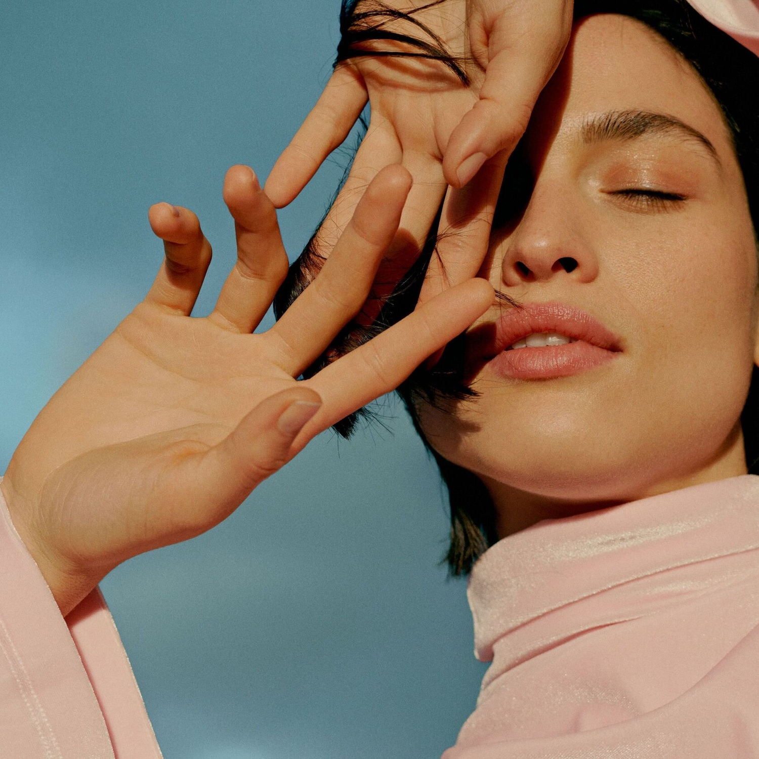 Une femme aux yeux clos, vêtue de rose, caresse son visage, créant une ambiance douce et rêveuse.