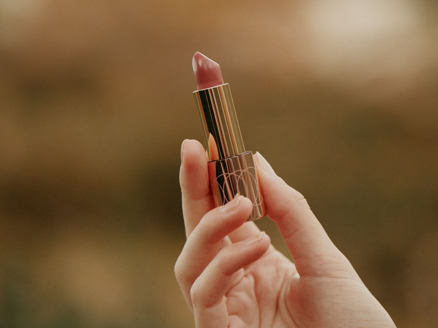 Voici un rouge à lèvres rose nude dans un élégant tube doré, tenu délicatement dans une main.