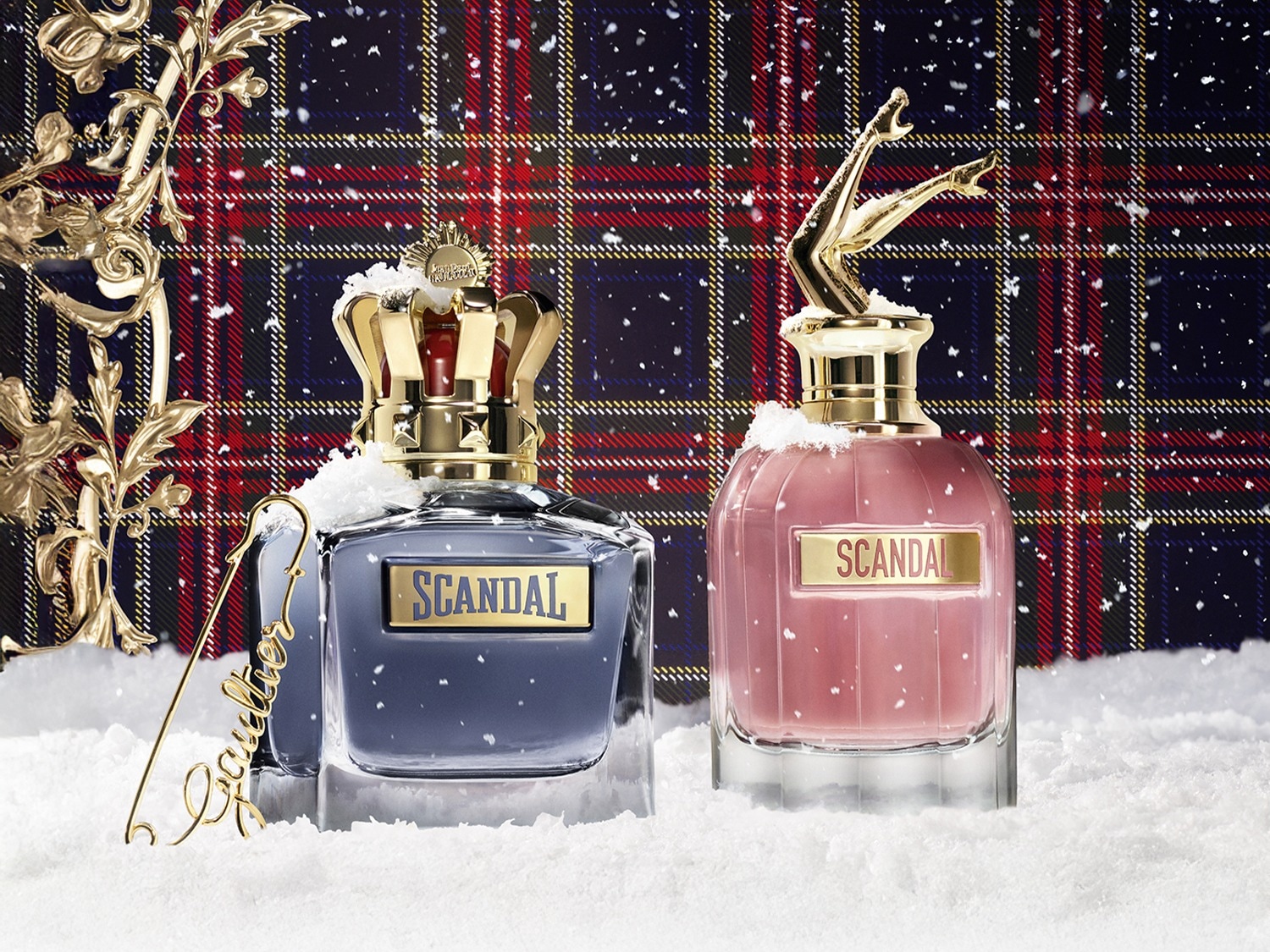 Twee flessen Scandal parfum, de ene blauw met een gouden kroon en de andere roze met gouden benen, staan in de sneeuw tegen een Schotse ruit achtergrond.