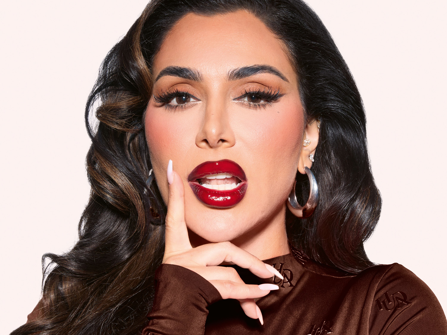 Nahaufnahme von Huda Kattan mit langen braunen Haaren, langen Wimpern und braunem Oberteil, die einen Finger an ihre glänzend roten Lippen hält.