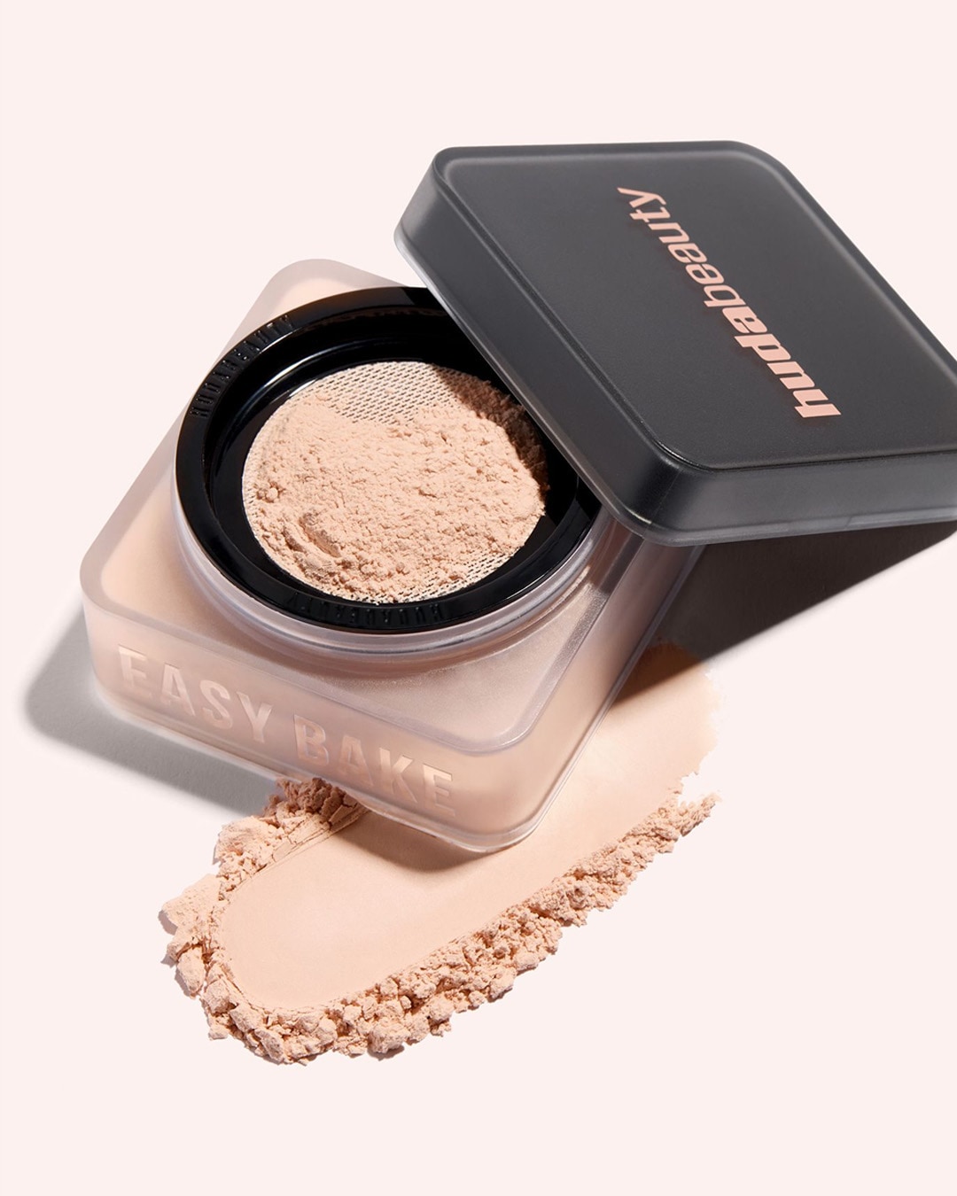 Huda Beauty Easy Bake Puder in einer durchsichtigen Dose mit offenem schwarzem Deckel, daneben eine Spur des losen Puders.
