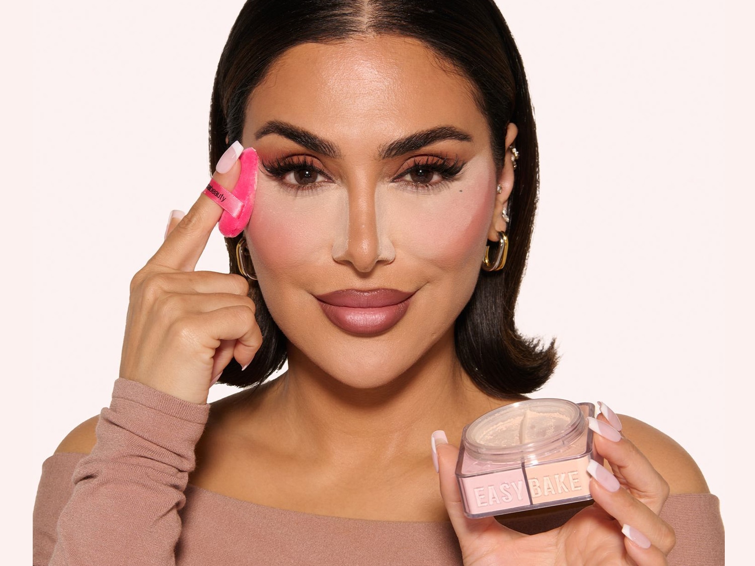 Huda Kattan hält in einer Hand einen rosa Make-up-Schwamm und in der anderen Hand ein transparentes Pudergefäß mit der Aufschrift EASY BAKE.