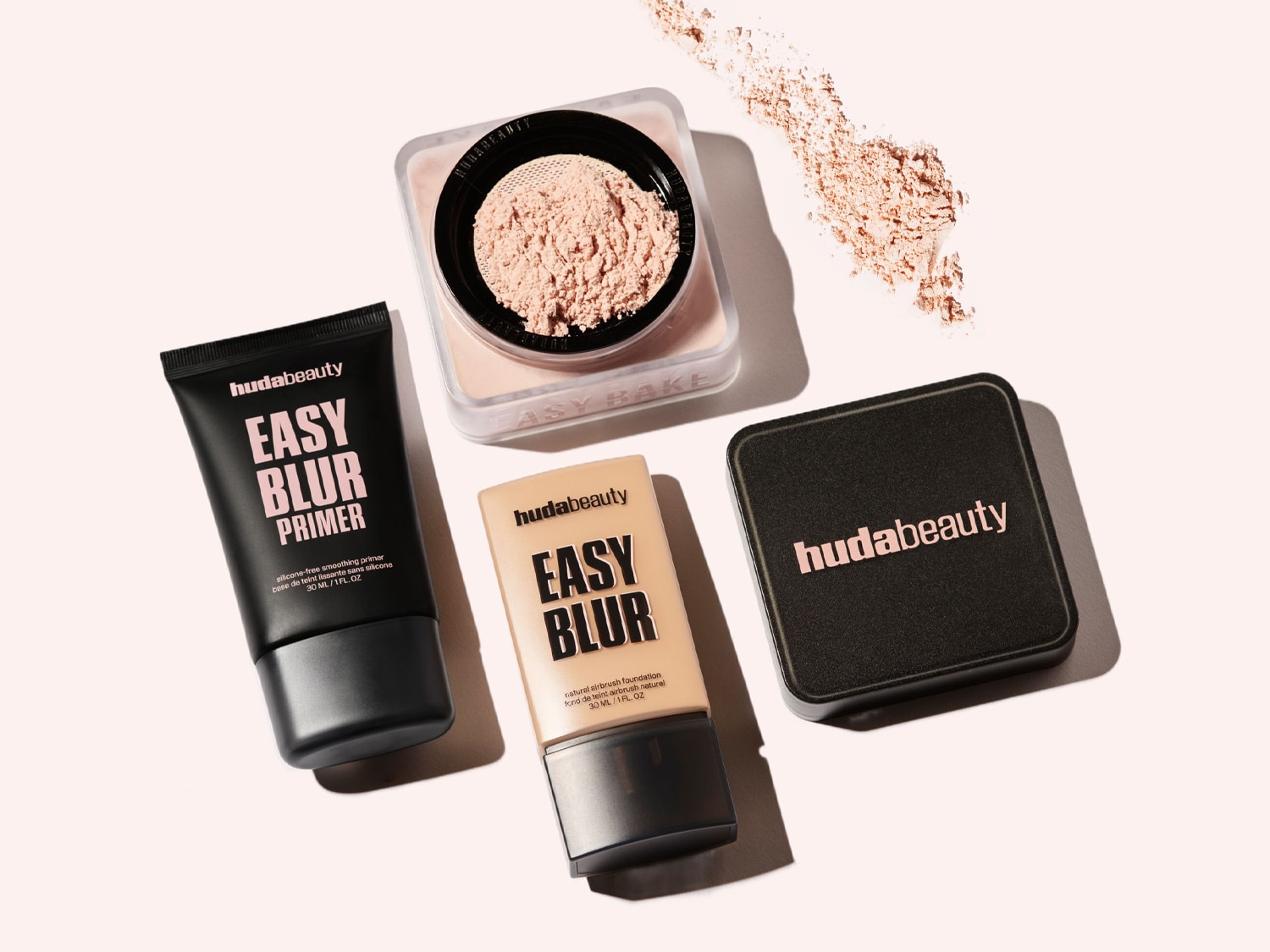 Huda Beauty Easy Blur Primer in einer schwarzen Tube, Easy Bake Puder in einer transparenten Dose mit geöffnetem Deckel und Easy Blur Foundation in einer beigen Tube sind auf beigem Untergrund drapiert.