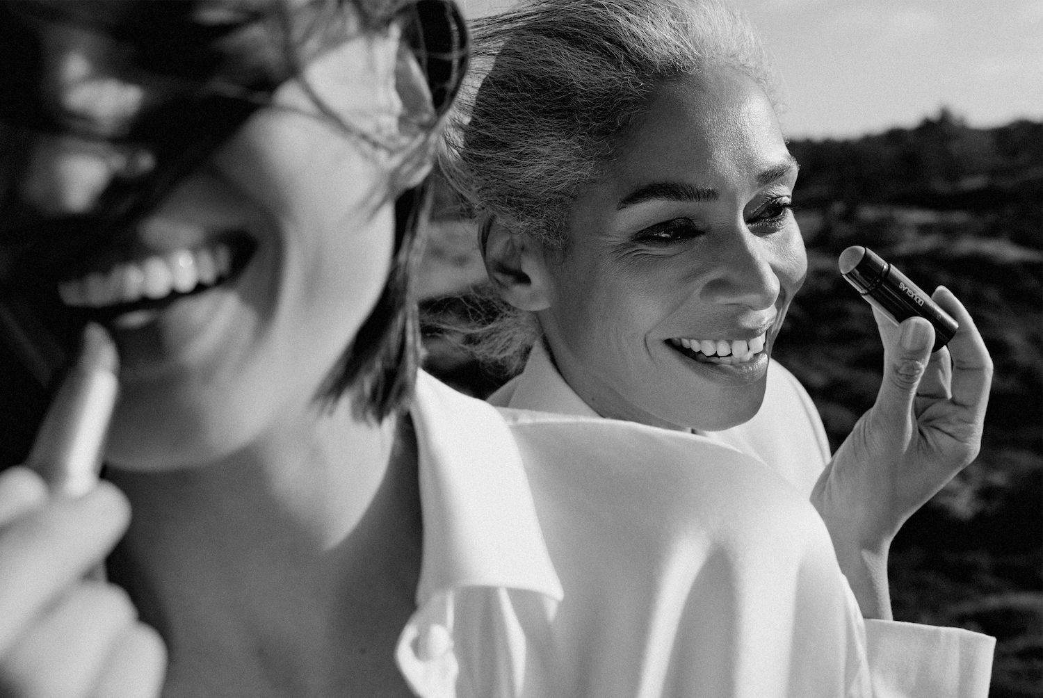 Zwei Frauen lächeln, eine hält einen Douglas Lippenstift in der Hand, während die andere einen Stift auf ihre Lippen aufträgt.