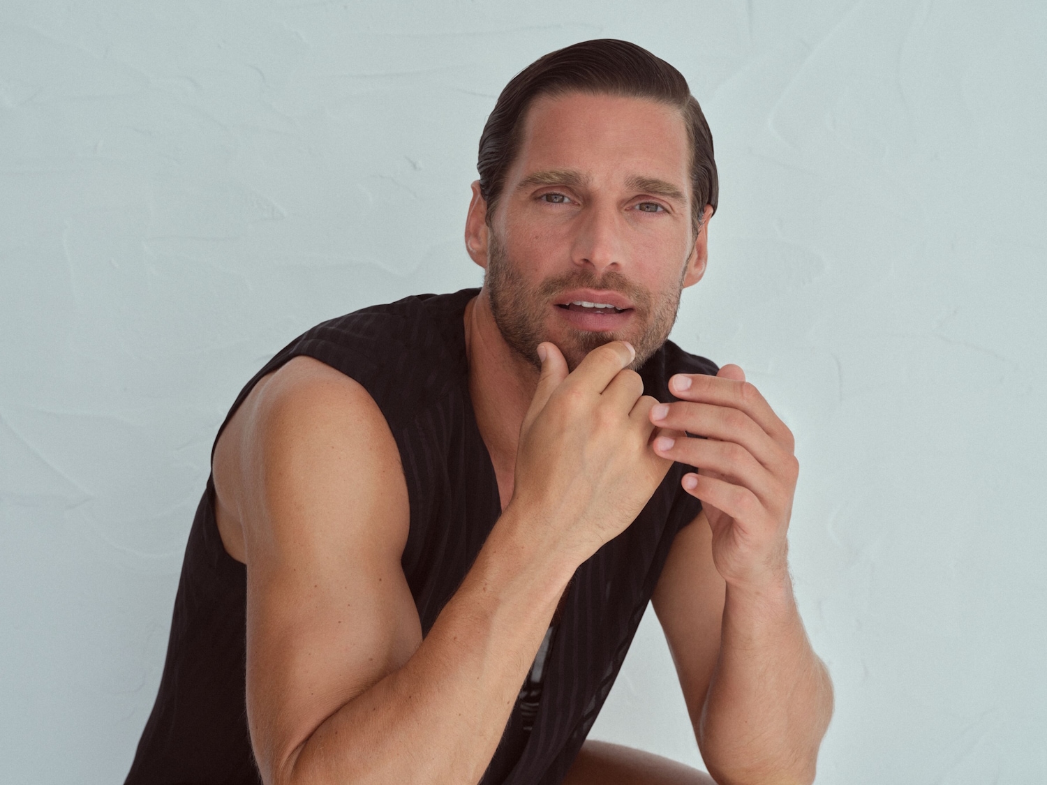Ein Mann mit geglätteten, dunklen Haaren und einem ärmellosen schwarzen Hemd posiert vor einer hellen Wand und berührt nachdenklich sein Kinn.