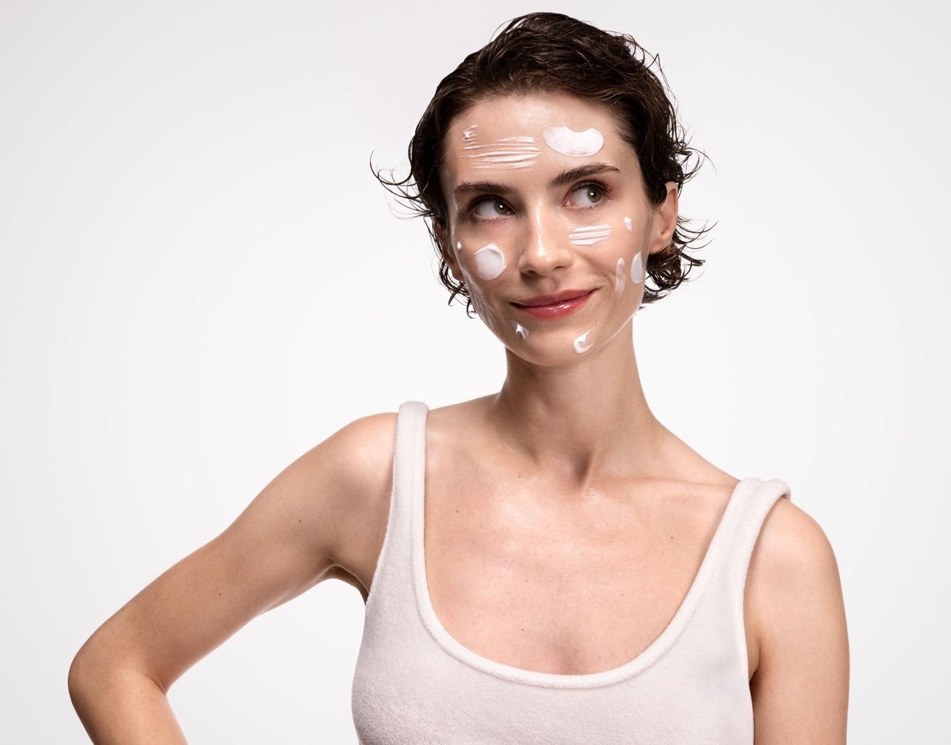 Eine Frau mit kurzen, dunklen Haaren und einem weißen Tanktop trägt weiße Creme auf ihrem Gesicht, während sie mit einem leichten Lächeln zur Seite schaut.