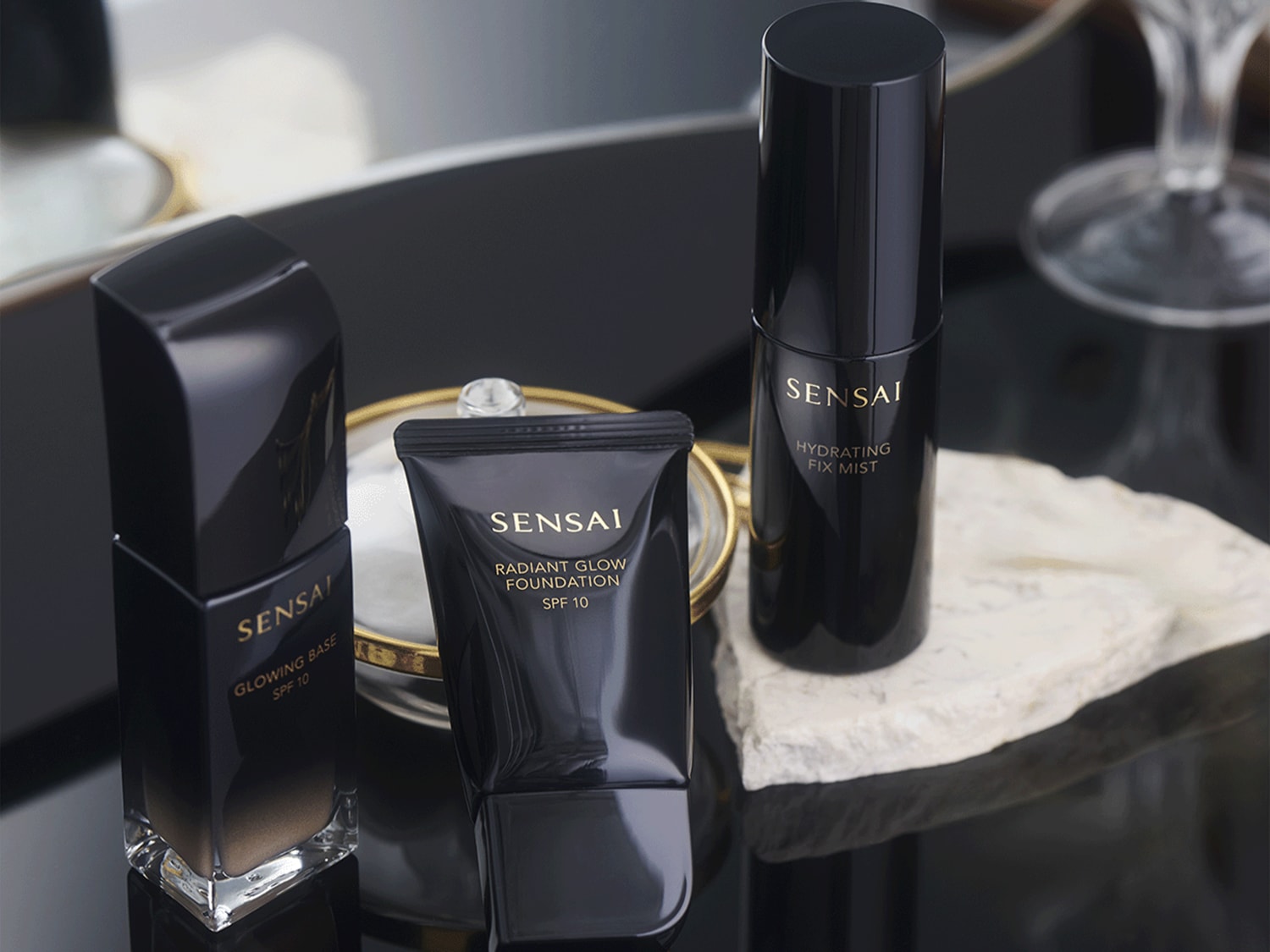 Elegancki zestaw kosmetyków Sensai w czarnych opakowaniach: baza rozświetlająca, podkład i mgiełka utrwalająca, zapewniające promienny wygląd.