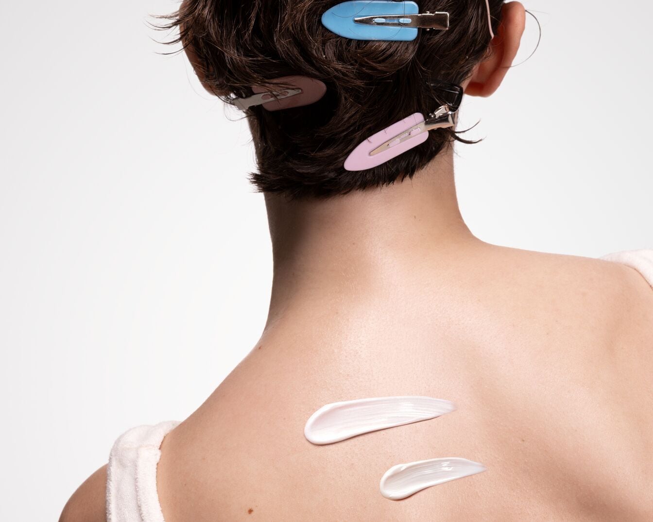 Nahaufnahme einer Frau mit Kurzhaarschnitt, deren Haare mit bunten Haarspangen zurückgesteckt sind, während auf ihrem Rücken zwei Kleckse weisser Creme liegen.