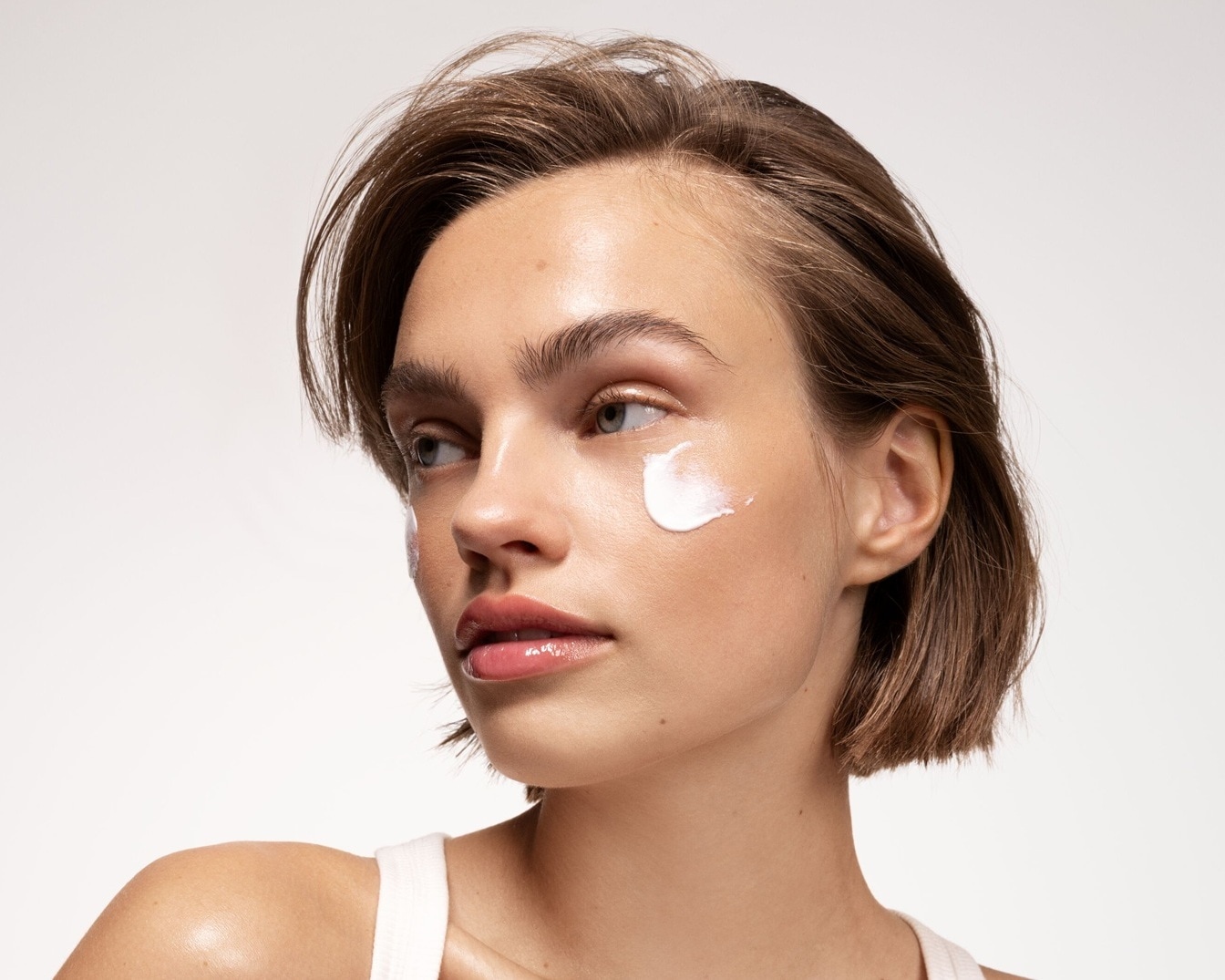 Nahaufnahme einer Frau mit kurzen braunen Haaren und einem weißen Top, die eine weiße Creme auf ihrer Wange unter dem Auge aufträgt.