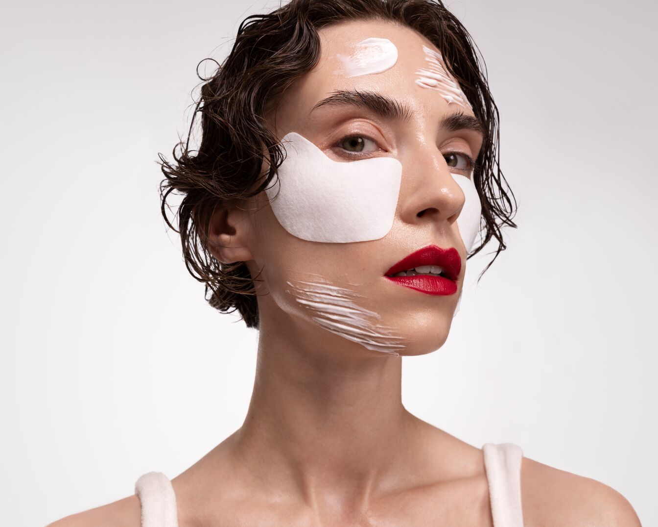 Nahaufnahme einer Frau mit roten Lippen, Augenpads und weißer Creme auf Gesicht und Stirn, die eine intensive Hautpflegeroutine zeigt.