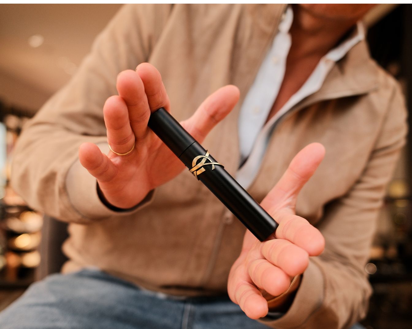 Een man toont een zwarte YSL mascara met gouden logo tussen zijn vingers, klaar om je wimpers te transformeren.