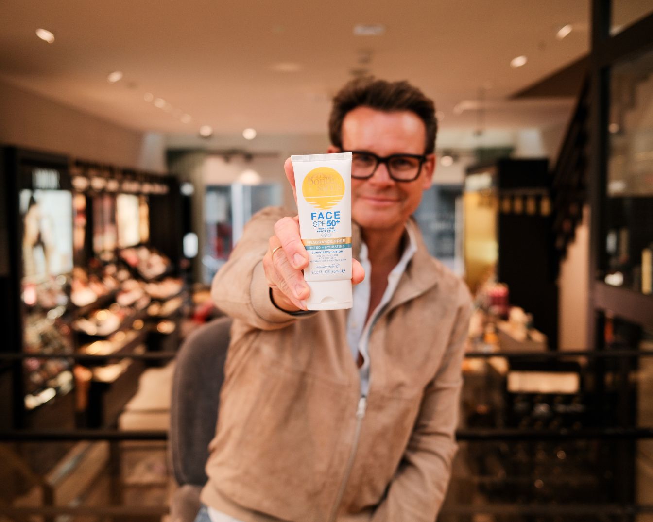 Een man met een bruin jasje en een bril houdt een witte tube zonnebrandcrème met SPF 50+ vast.