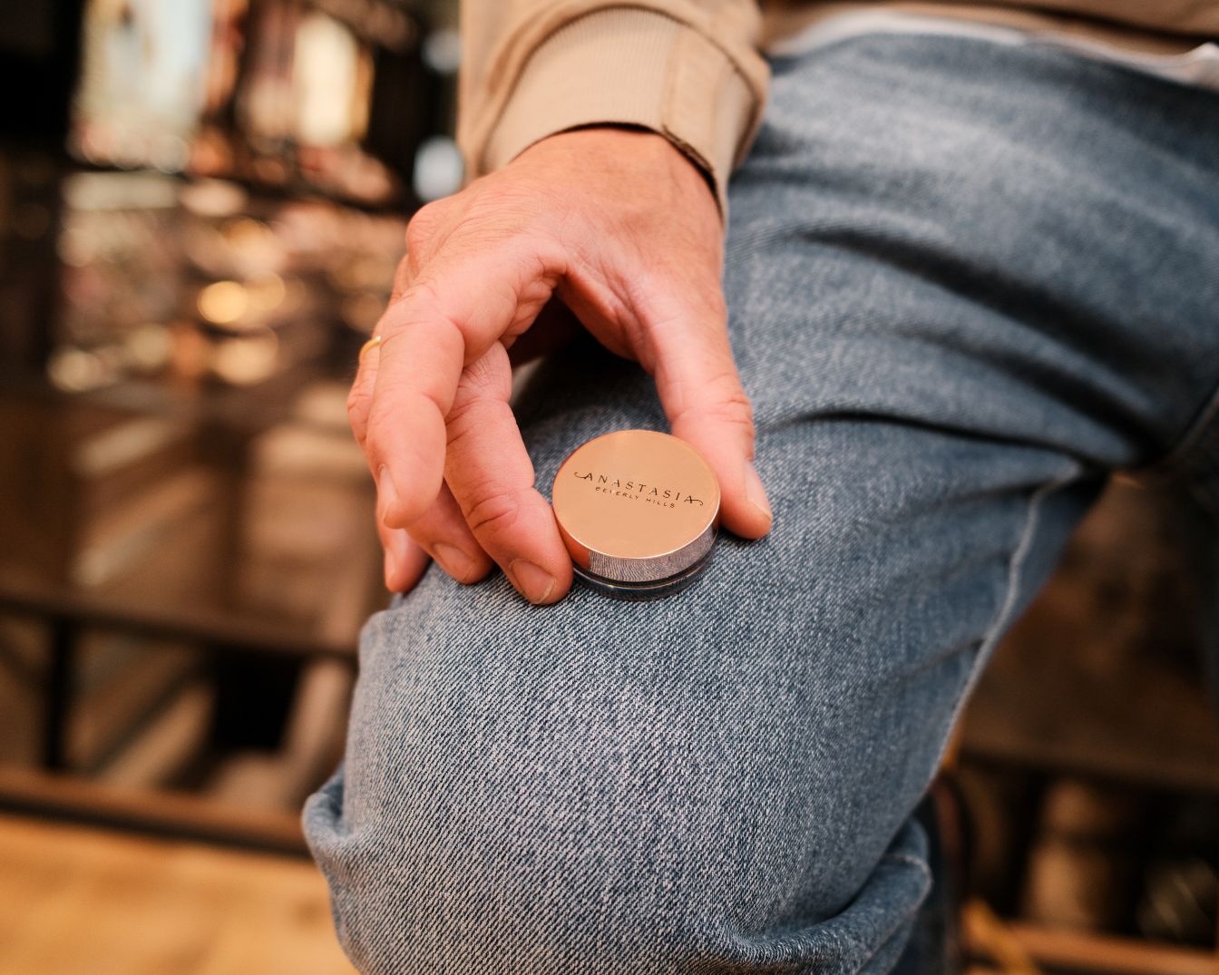 Een hand houdt een ronde, goudkleurige Anastasia Beverly Hills make-updoos vast tegen een blauwe jeans.