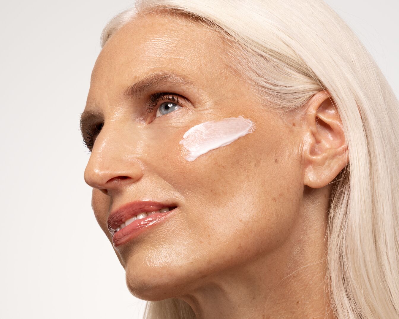 Nahaufnahme einer Frau mit weißblondem Haar und einem Klecks weißer Creme auf der Wange, die die Feuchtigkeitspflege für reife Haut demonstriert.