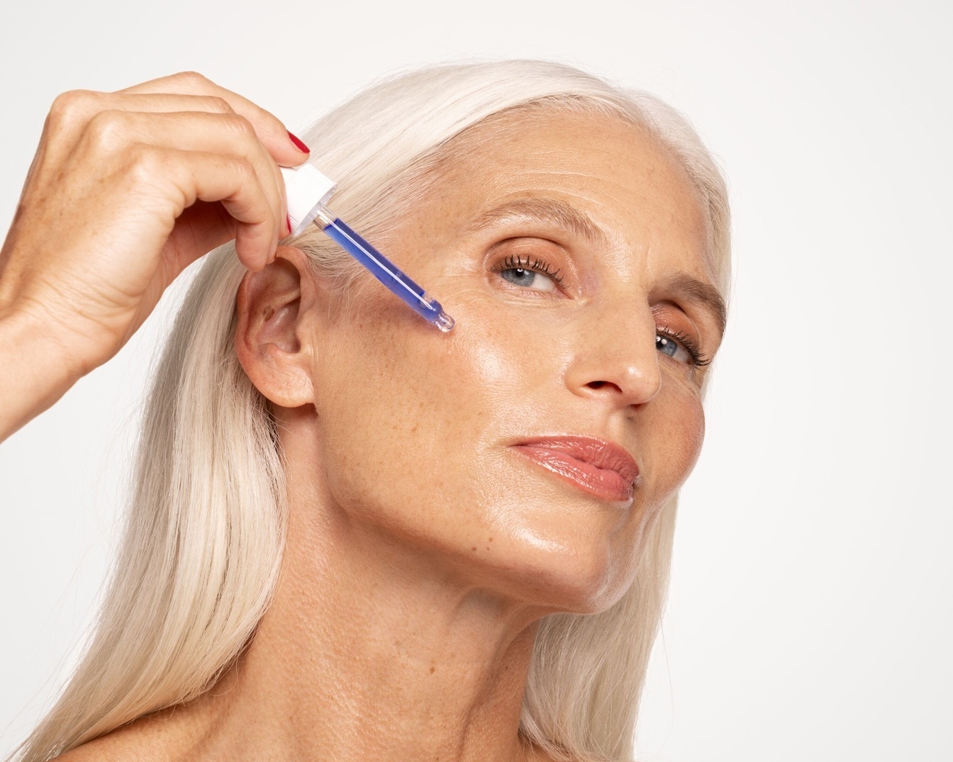 Eine Frau mit silberblondem Haar trägt ein blaues Serum mit einer Pipette auf ihr Gesicht auf, um ihre Haut zu pflegen.