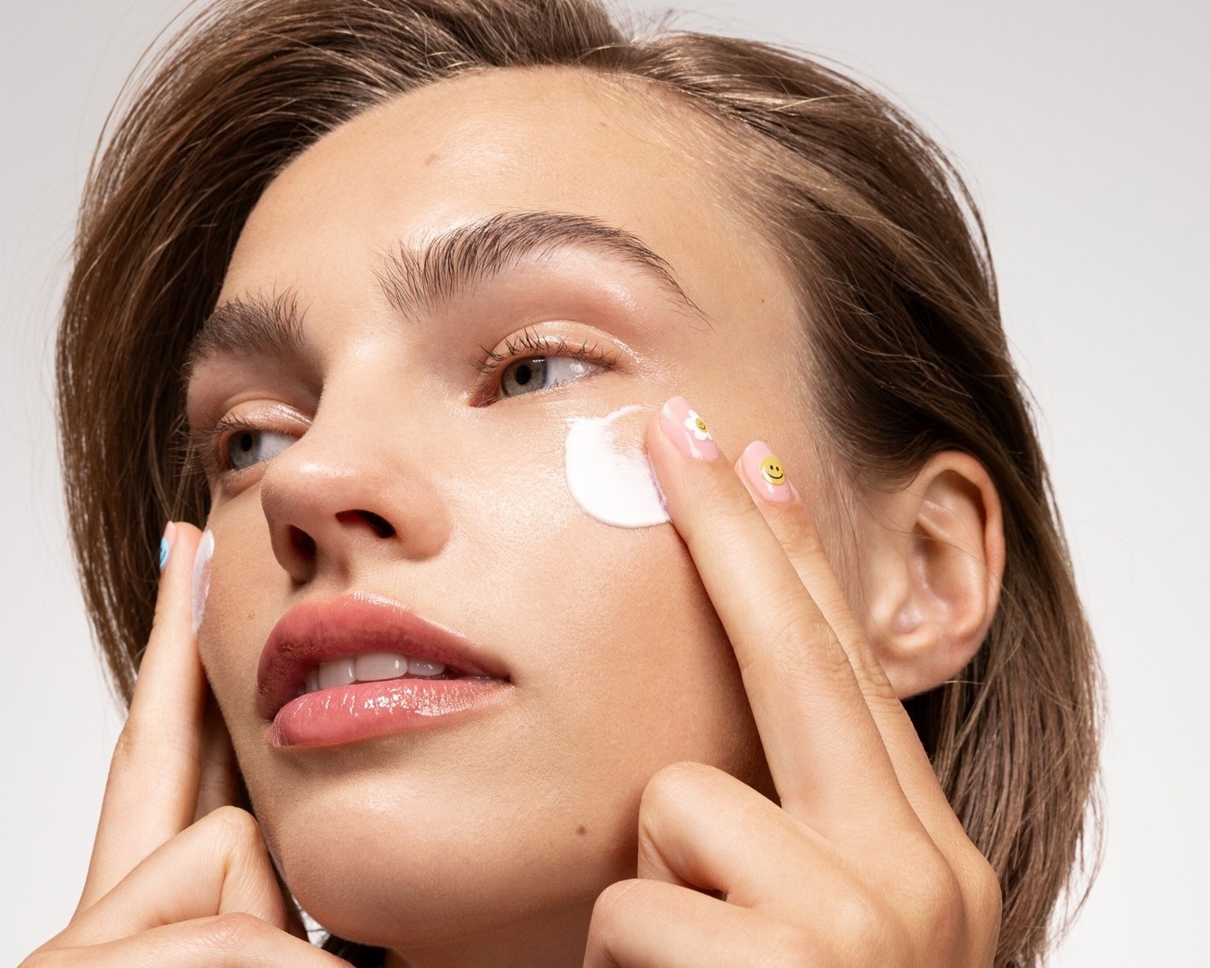 Eine Frau mit hellbraunem Haar trägt eine weiße Creme auf ihre Wange auf, wobei ihre Finger mit Nagellack mit Smiley- und Blumenmotiven verziert sind.