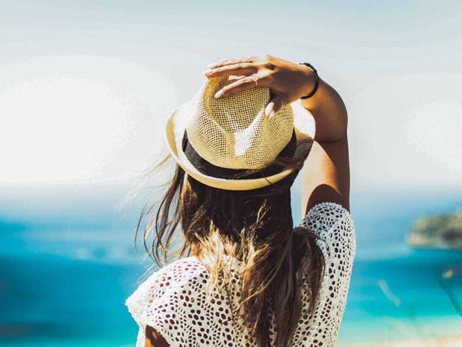 Una donna con un cappello di paglia guarda l'orizzonte sul mare azzurro, indossando un top bianco traforato.