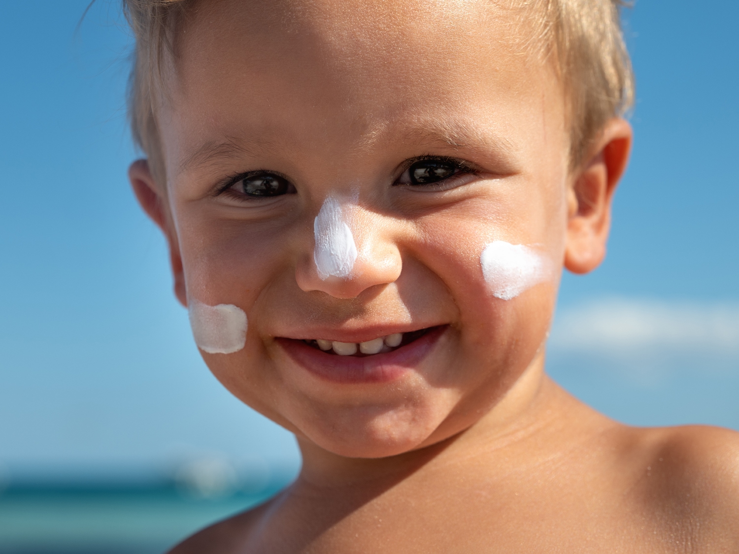 Primo piano di un bambino sorridente con crema solare bianca applicata su naso e guance, sullo sfondo un cielo azzurro.